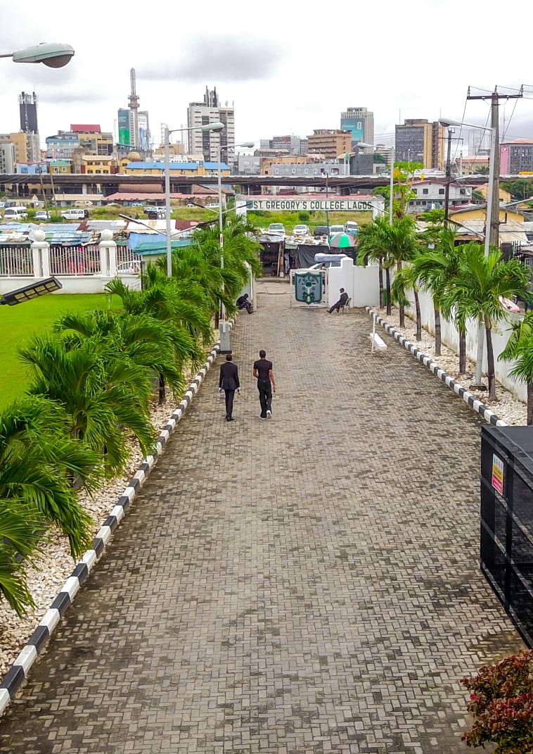 Students walking the palm-lined entrance drive with the Lagos skyline beyond